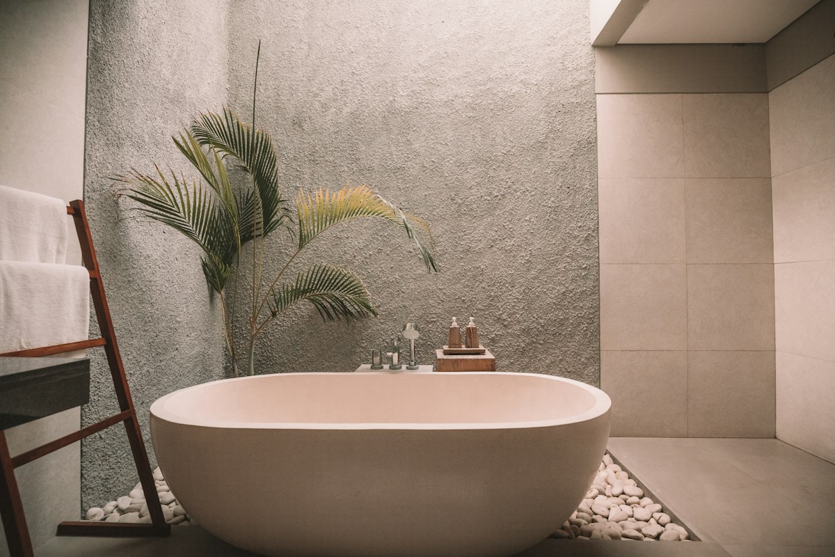 Freestanding oval soaking tub with river stones, palm plant, and towel ladder in stone bathroom.